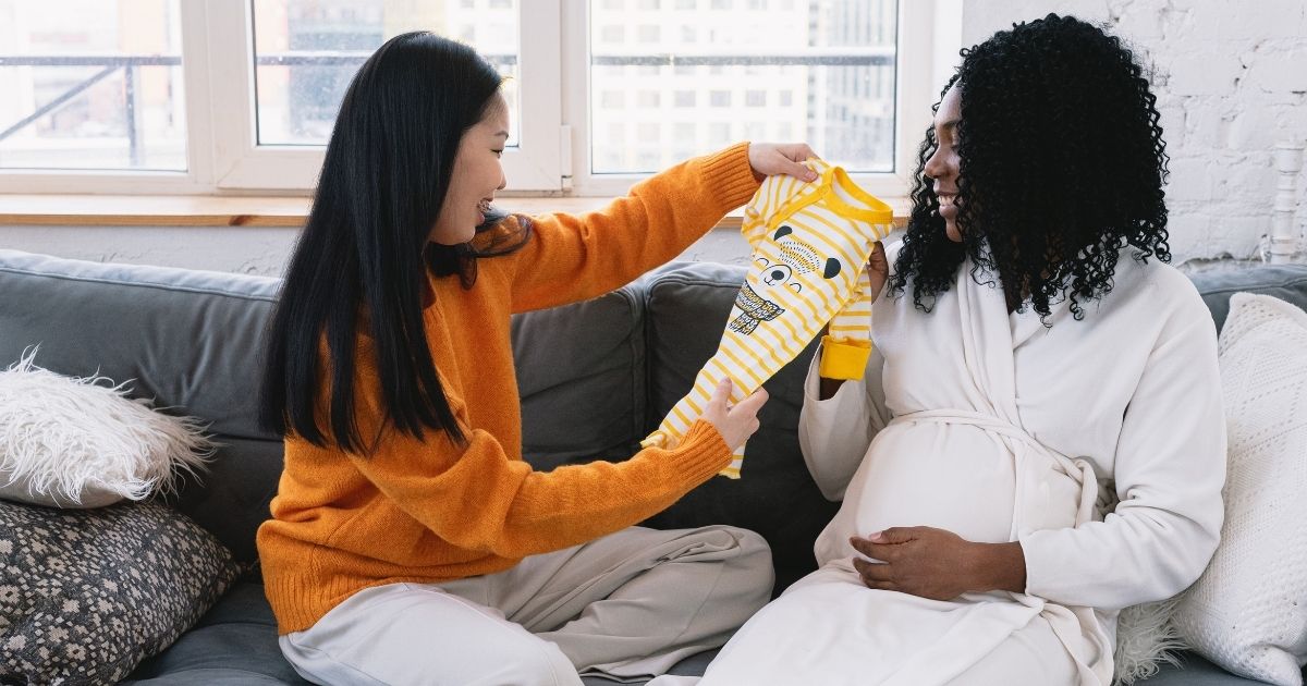 Photo of a lesbian couple, one an Asian woman wearing an orange sweater holding up a yellow printed onesie, and her partner a pregnant Black woman with curly shoulder length hair, white robe.