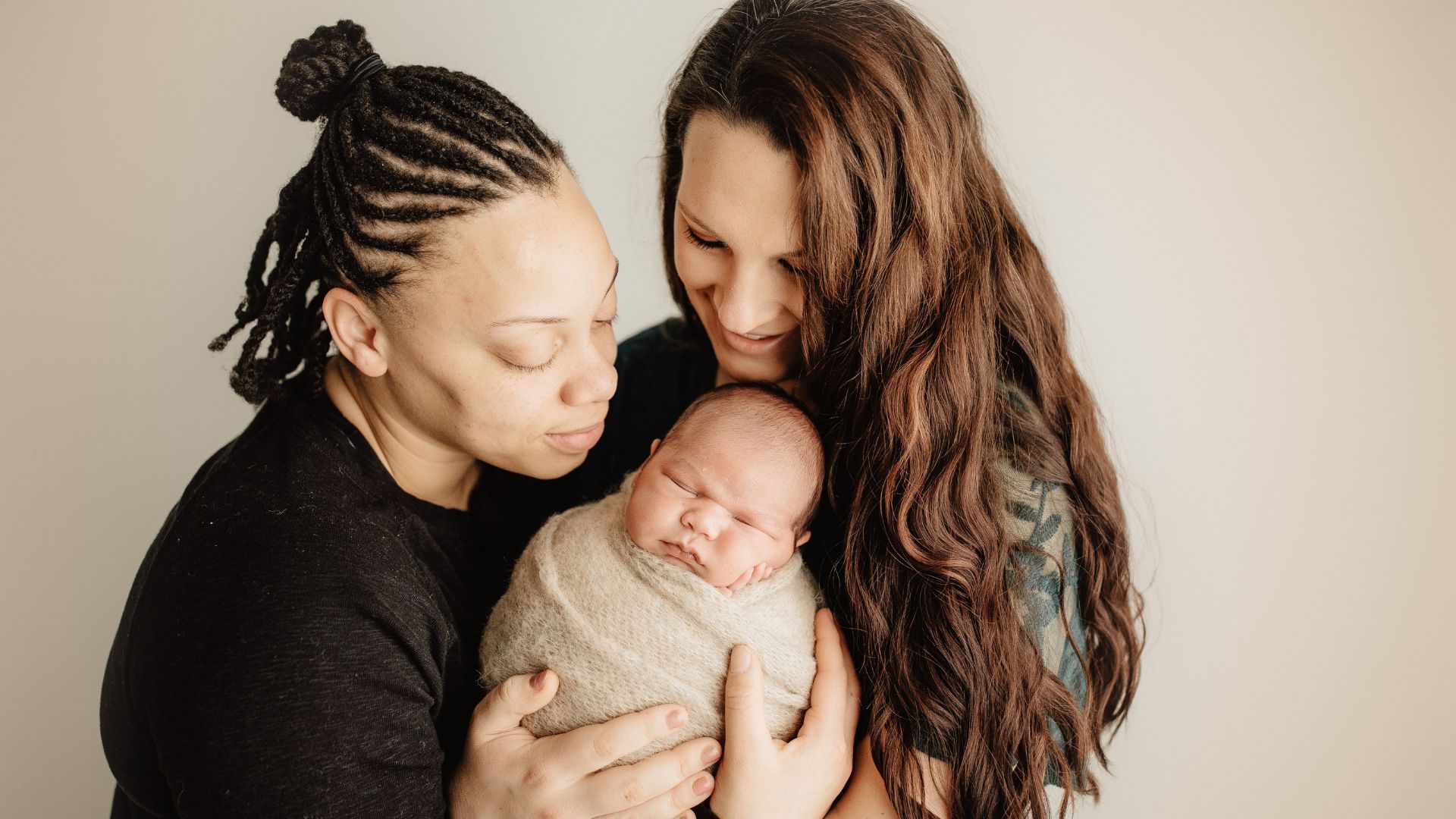 Photo of an interracial couple in the LGBTQIA+ community, holding their new baby.