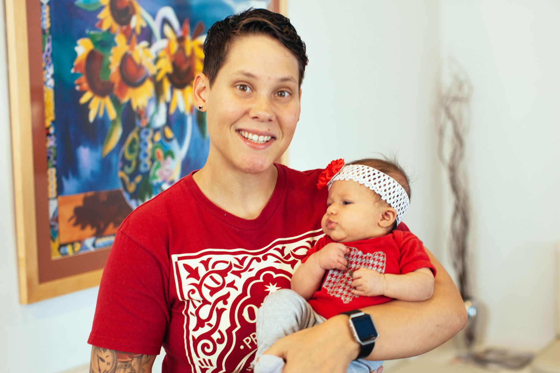 Photograph of a person in a red printed graphic t-shirt with short, clean cut hair, smiling, holding a baby who looks around 3 months old wearing a matching red onesie and floral headband. In the background is a painting of sunflowers in a gold frame.