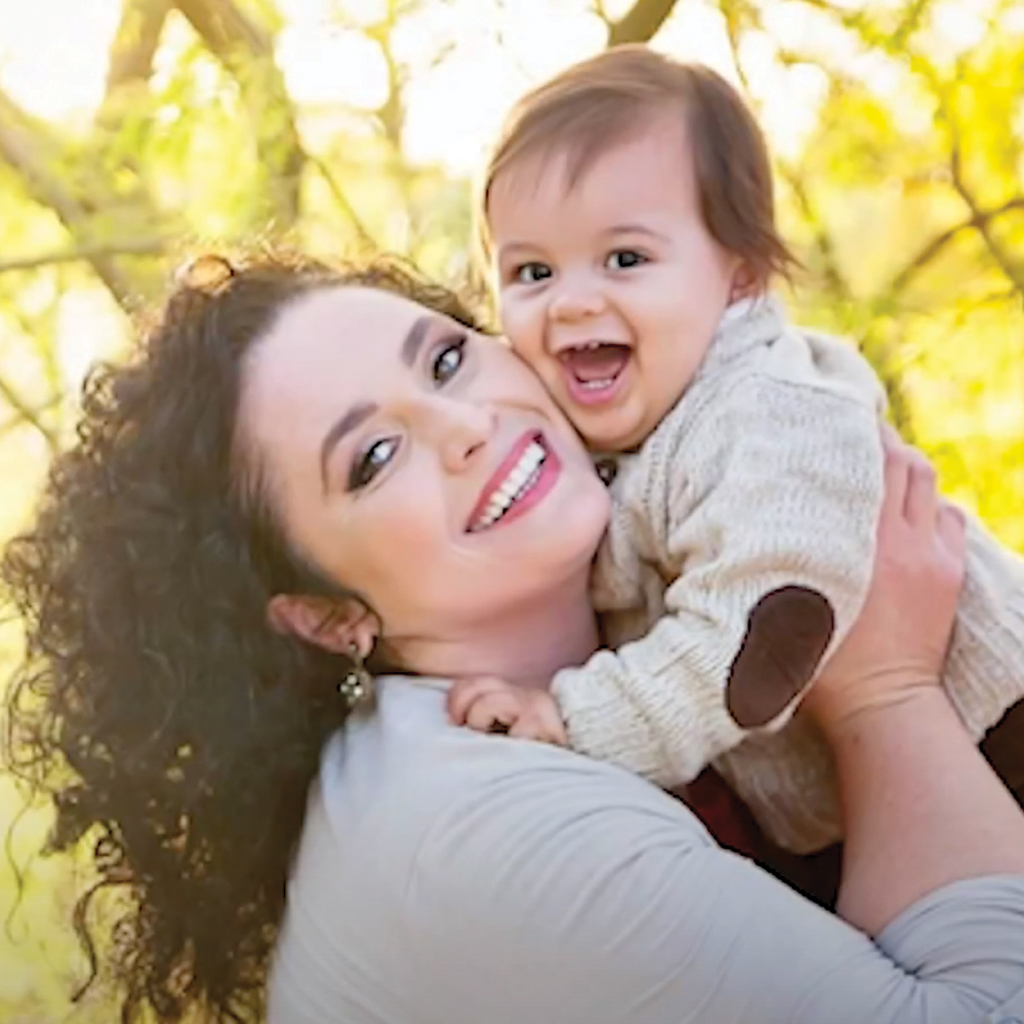 Photograph of sunny light speckled yellow leaf trees as a blurry background with a smiling woman named Shyla who has curly brown hair, a beige sweater, and a large smile, holding her baby who looks to be about one year old, with a brown head of full hair, wearing a sweater with elbow patches.
