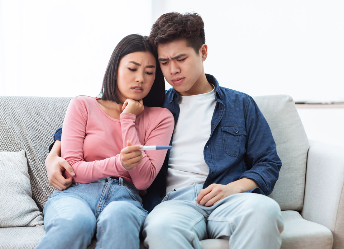 Frustrated Asian couple sitting on the couch looking at a pregnancy test.